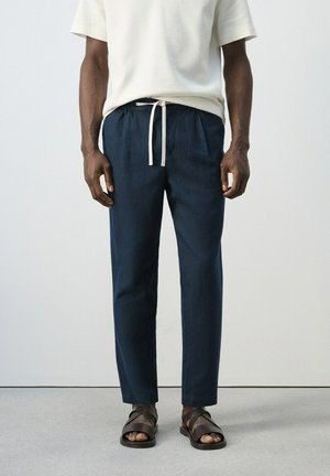 Hombre vestido con pantalones azul marino con cordón, camisa blanca de manga corta y sandalias de cuero marrón, de pie contra un fondo liso.