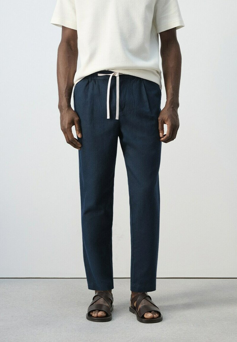 Man draagt marineblauwe broek met trekkoord, wit kort mouwen shirt en bruine leren sandalen, staande tegen een effen achtergrond.