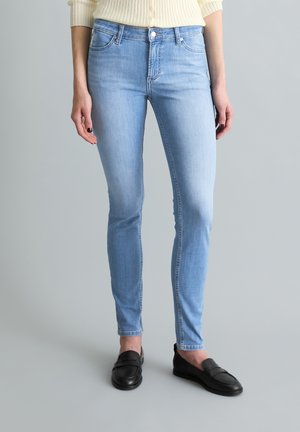 Frau, die hellblaue Skinny-Jeans und schwarze Loafers trägt, steht vor einem schlichten grauen Hintergrund.