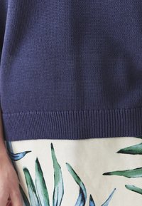 Maglione lavorato a maglia blu navy con orlo a coste, abbinato a un tessuto crema con motivi di foglie verdi, mostrando contrasto di texture e colore.