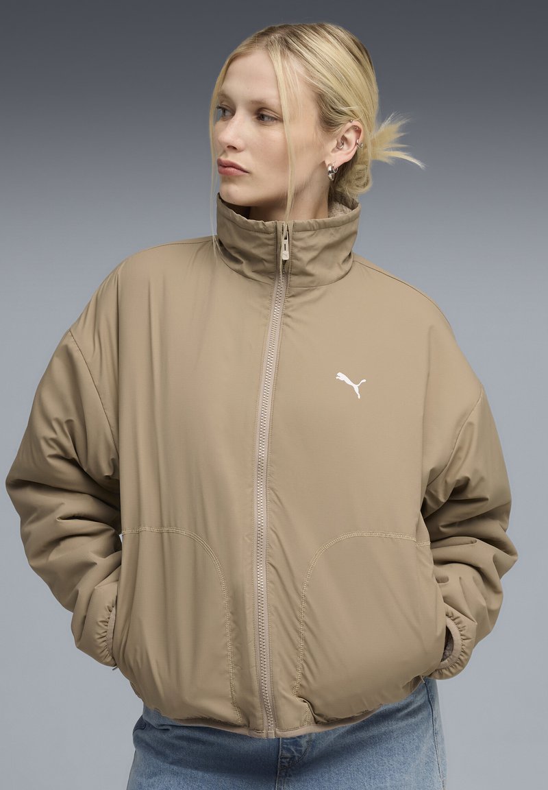 Giacca imbottita beige con colletto alto, chiusura con zip e logo PUMA. Presenta grandi tasche laterali e polsini elasticizzati. Tessuto morbido e leggero.