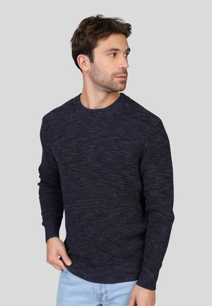 Pull en tricot bleu marine avec un col rond, des manches longues et un tissu texturé. Porté avec un jean bleu clair. Design sobre.