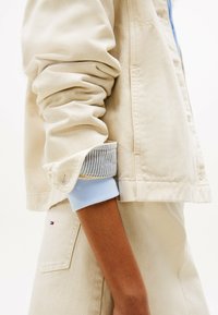 Chaqueta de mezclilla beige con costuras visibles, mangas enrolladas que muestran puños de rayas en azul claro. Presenta un pequeño logotipo en el bolsillo trasero.