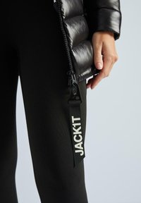 Juodas pūkinis švarkas su blizgesiu, atsegta, kad būtų matoma tekstūruota juoda legingo kelnės, ant kurios peties yra balta "JACK1T" etiketė.