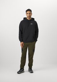 Hoodie noir avec une poche avant, associé à un pantalon kaki avec des accents noirs et rouges, et des chaussures de sport grises.