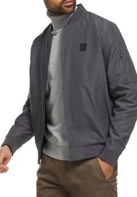 Grijze bomberjack met een ritssluiting aan de voorkant en ribgebreide manchetten. Heeft een ritszak op de mouw en een logo-applicatie op de linkerborst.