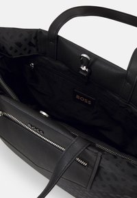 Sac à main en cuir noir avec un extérieur texturé, fermeture éclair et poche intérieure. Il présente une étiquette de marque et des poignées robustes pour un transport facile.