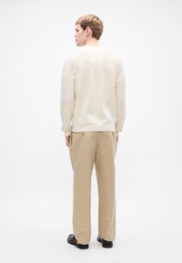 Giovane uomo di spalle, che indossa una camicia a maniche lunghe color crema, pantaloni beige con tasche posteriori e scarpe nere su uno sfondo bianco uniforme.