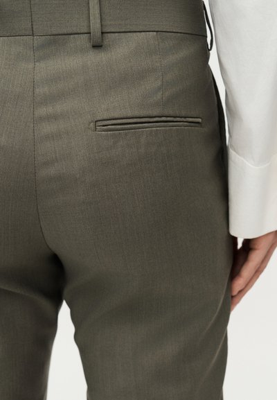 Κοντινή λήψη προσώπου που φοράει ελιάς πράσινο ραμμένο παντελόνι με πίσω τσέπη με άνοιγμα και λευκό μακρυμάνικο πουκάμισο.