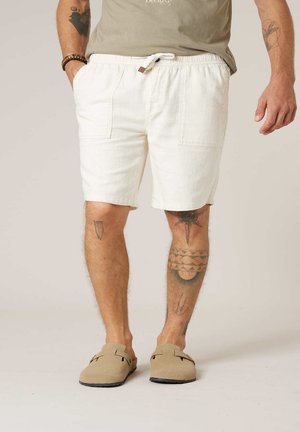 Man draagt witte shorts met trekkoord, beige instapschoenen en een groen shirt, toont tatoeages op armen en benen tegen een effen achtergrond.