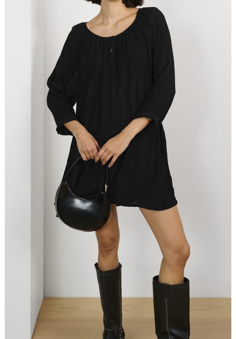 Robe noire, à mi-longueur, avec des manches trois-quarts et un col arrondi. Associée à un sac à main noir rond et des bottes noires hautes.