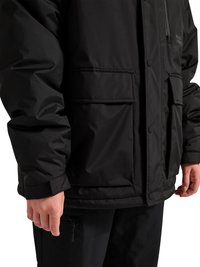 Mann trägt schwarze isolierte Jacke mit Klappentaschen und Reißverschlusstasche auf der Brust, kombiniert mit schwarzen Hosen.