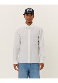Camicia bianca di lino con maniche lunghe e chiusura a bottoni, abbinata a jeans in denim azzurri. Modello che indossa un cappellino da baseball nero.