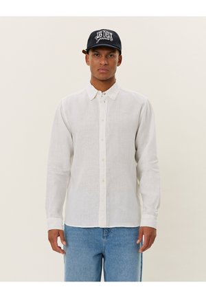 Camicia bianca di lino con maniche lunghe e chiusura a bottoni, abbinata a jeans in denim azzurri. Modello che indossa un cappellino da baseball nero.
