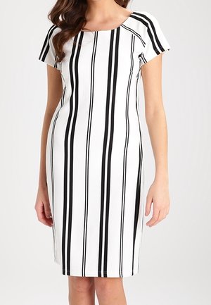 Kvinde iført en knælængde hvid kjole med korte ærmer og sorte lodrette striber, stående mod en ensartet baggrund.