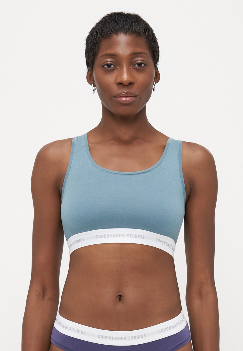 Blå bomulls-bralette med scoop-neck og hvit elastisk band med "COPENHAGEN STUDIOS" logo. Glatt tekstur, tettsittende design.
