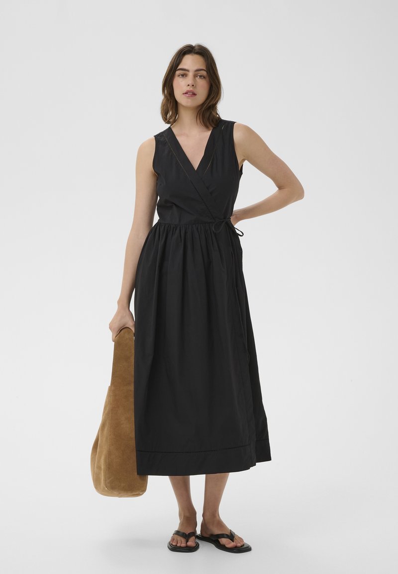 Femme debout, portant une robe portefeuille noire sans manches et des sandales noires, tenant un grand sac fourre-tout en daim marron dans une main.