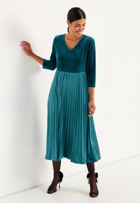 Türkises Samtkleid mit plissiertem Rock, V-Ausschnitt und Dreiviertelärmeln. Mit tailliertem Oberteil und fließenden Falten bis zur Mitte der Waden.