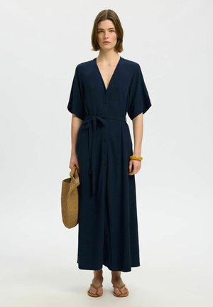 Frau trägt ein dunkelblaues Kleid mit Knopfleiste und Gürtel, hält eine geflochtene Tasche, trägt beige Sandalen und ein gelbes Armband, steht vor weißem Hintergrund.