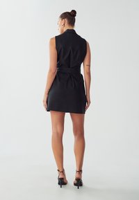 Vestito nero senza maniche con un colletto strutturato e vita cinturata. Tessuto morbido, lunghezza mini, indossato con tacchi neri con cinturini.