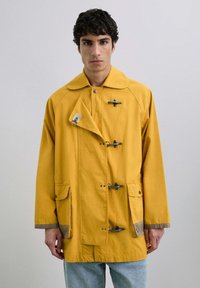 Chaqueta amarilla de gran tamaño hecha de algodón con un diseño de doble botonadura, toggles de madera y dos bolsillos frontales; presenta acentos grises en los puños.