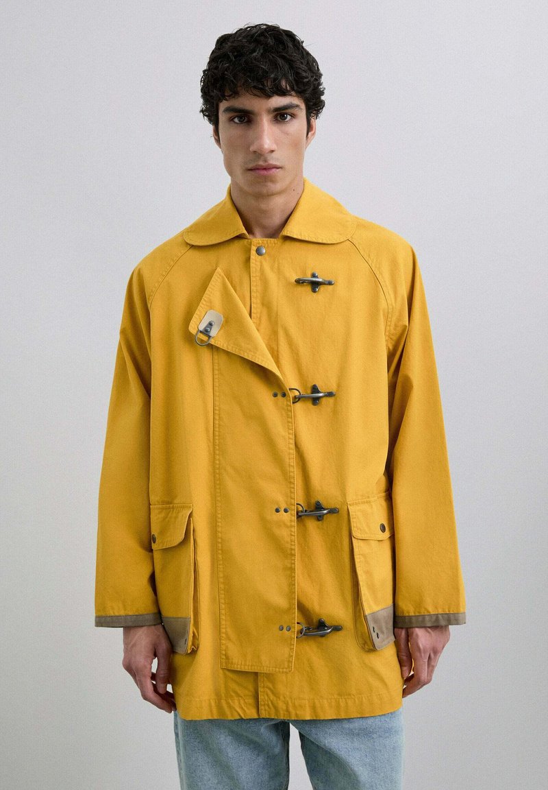 Chaqueta amarilla de gran tamaño hecha de algodón con un diseño de doble botonadura, toggles de madera y dos bolsillos frontales; presenta acentos grises en los puños.