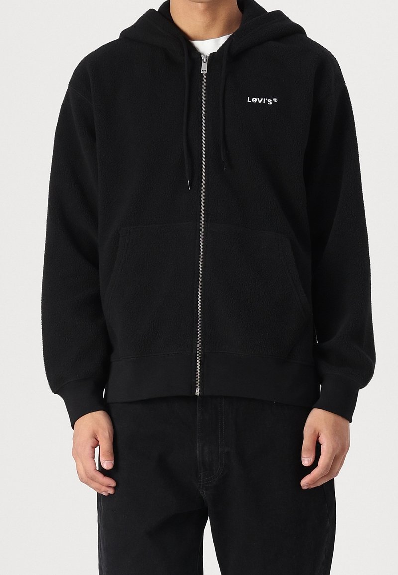 Man i svart fleece zip-up hoodie från Levi's med framfickor och dragsko på huvan, tillsammans med svarta byxor.