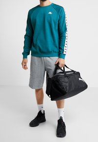 Muž v tyrkysovém svetru, šedých šortkách, černých teniskách a bílých ponožkách drží černou sportovní tašku Puma proti jednobarevnému pozadí.