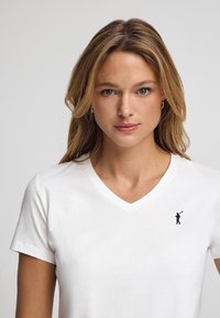 Hvid V-hals t-shirt lavet af bomuld, med et lille marineblåt grafik af en golfspiller på venstre brystområde. Korte ærmer og standard pasform.