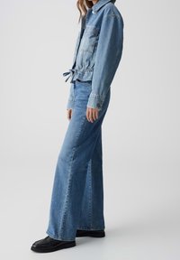 Světle modrá džínová bunda s stahovací šňůrkou v pase, náprsními kapsami a dlouhými rukávy, kombinovaná s širšími džíny ve stejné barvě a černými botami.