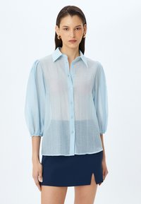 Camisa de botones azul claro y semitransparente con tejido texturizado y mangas abullonadas, que presenta un cuello con solapa y un ajuste ligeramente holgado.