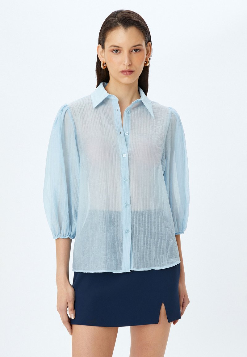 Camisa de botones azul claro y semitransparente con tejido texturizado y mangas abullonadas, que presenta un cuello con solapa y un ajuste ligeramente holgado.