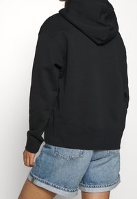 Zwarte hoodie met een grote capuchon, gemaakt van een zachte katoenmix. Heeft een relaxte pasvorm en ribgebreide manchetten. Gemaakt in combinatie met lichte denimshorts.