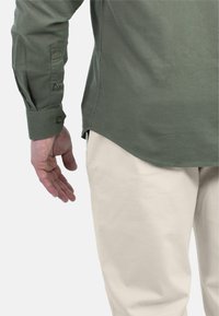 Olijfgroen langemouwenshirt met knoopmanchetten, gecombineerd met crèmekleurige broek. Het shirt heeft een ronde zoom en een gladde textuur.