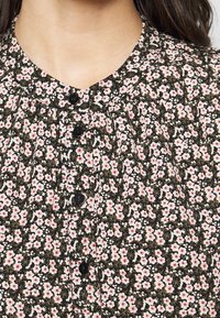 Květinová blůza z tmavé látky s malými růžovými a bílými květinami. Má kulatý límec a černé knoflíky vpředu se sklady.