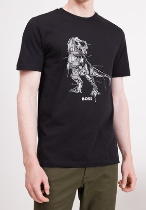 Schwarzes T-Shirt mit weißem Leiterplattendesign, das einen Löwen und das Wort "BOSS" darunter bildet, getragen von einer Person mit olivgrüner Hose.