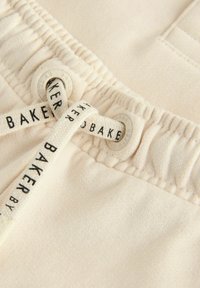 Tissu beige clair avec une taille élastique, comportant deux œillets ronds et un cordon de serrage étiqueté "BAKER". Texture lisse avec des bords froncés.