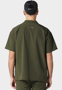 Grønn kortermet skjorte laget av et lett stoff; har en rett passform med en subtil preget logo på baksiden.