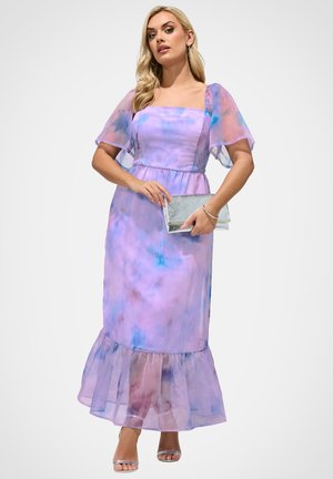 Frau trägt ein maxikleid in Lavendel- und Blautönen mit Batikmuster, mit flatternden Ärmeln, hält eine silberne Clutch und trägt silberne offene Sandaletten.