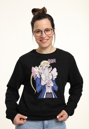 Lächelnde Frau mit Brille und Dutt trägt einen schwarzen Pullover mit einem schockierten Yu-Gi-Oh! GX-Charakter, der ein Tuch festhält.