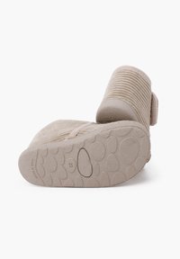 Beige slipper met een bovenkant van geprofileerde stof, een verstelbare band en een rubberen zool met een gestippeld patroon voor grip. Subtiele stikdetails zichtbaar.