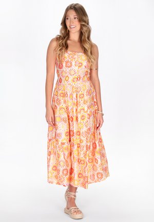 Kvinde med langt, bølget hår iført en stropløs midi-kjole med orange, lyserøde og gule blomster og beige snøresandaler, stående på hvid baggrund.