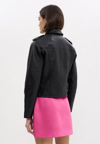 Donna di spalle che indossa una giacca di pelle nera e una gonna rosa brillante su uno sfondo bianco uniforme.