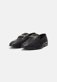 Black loafers made of textured leather, featuring a decorative gold-colored bar across the front and a sleek, pointed toe design.