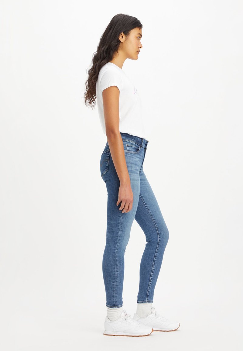 Jean skinny bleu en denim, dotés d'une taille haute, avec une silhouette ajustée et un léger délavage, associés à un t-shirt blanc à manches courtes et des baskets blanches.