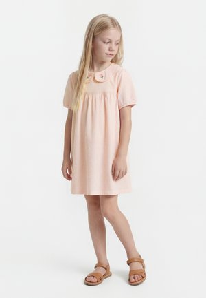 Junges Mädchen mit langen blonden Haaren, das ein hellrosa Kleid mit besticktem Kragen und braune Sandalen trägt, steht und schaut nach unten.