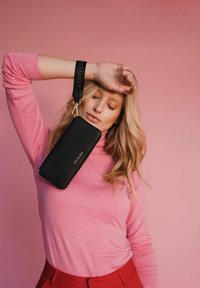 Zwarte textuur portemonnee met een gouden rits, bevestigd aan een zwarte band met "HEY MARLY" gegraveerd, gedragen door een persoon in een roze top.