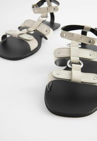 Sandali in ecopelle beige con suole nere piatte, dotati di molteplici cinturini e dettagli metallici per supporto e vestibilità regolabile.
