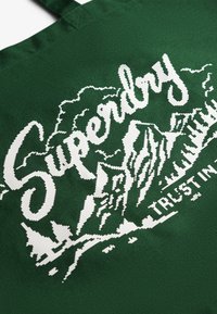 Groene canvas draagtas met witte grafische tekst "Superdry" en een ontwerp van een berglandschap, gemaakt van stevige stof en met dubbele schouderbanden.