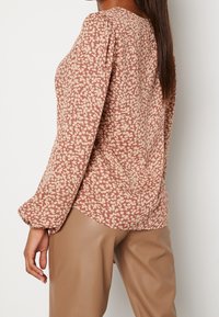 Blusa a maniche lunghe di un rosa tenue con piccolo motivo floreale color crema, presenta spalle arricciate e un orlo curvo, abbinata a pantaloni beige lucidi.